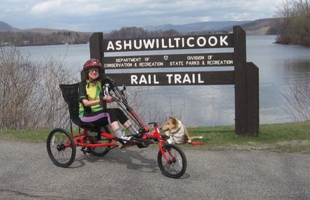 Person in recumbant adapted bicycle in fron to Ashuwillticook Rail Trail sign